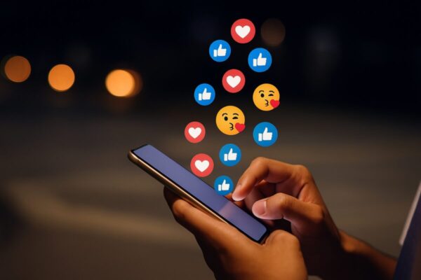 Mano usando smartphone con íconos flotantes de redes sociales como likes y emojis, representando el impacto en la autoestima adolescente
