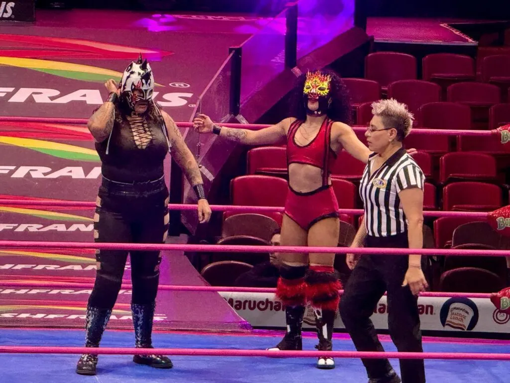 Dos luchadoras profesionales enmascaradas aparecen junto a una réferi dentro del cuadrilátero de la Arena México.