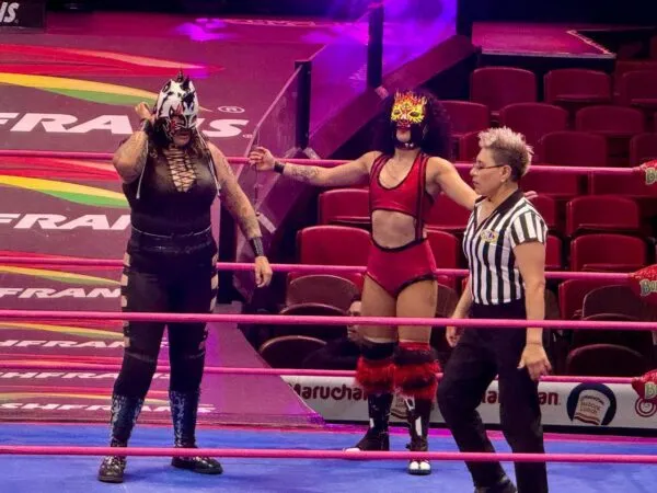 Dos luchadoras profesionales enmascaradas aparecen junto a una réferi dentro del cuadrilátero de la Arena México.